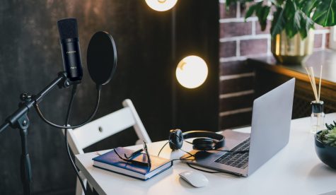 Items for recording podcast: professional microphone, earphones and laptop on white table in cozy home studio with black walls and lots of plants.
